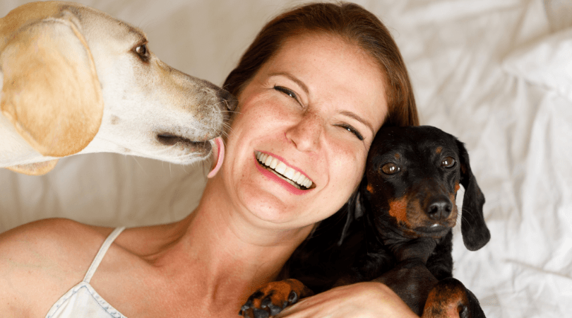 A lady getting a kiss from one dog while another dog looks at her on her bed.